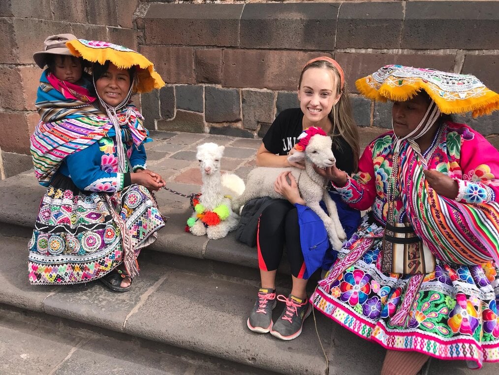 A girl holding a baby lamb sits with two women wearing traditional Peruvian dress.